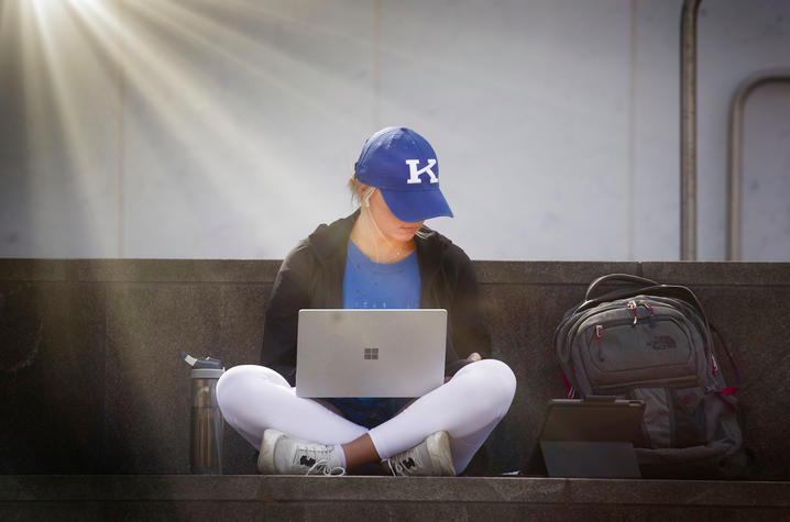 A female student in a UK baseball cap works on a laptop outdoors in the sunlight.
