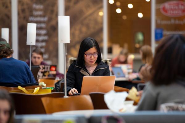 A student glances at their laptop with a look of concentration as they sit at a table underneath a white lamp. Students surround them at other tables in various stages of lounging and concentration.