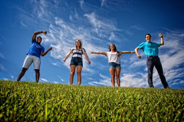 Four students stand together in a field of grass amidst a backdrop of a lightly cloudy blue sky. The four students stand in a line and move their arms to spell out the word 'CATS.' 