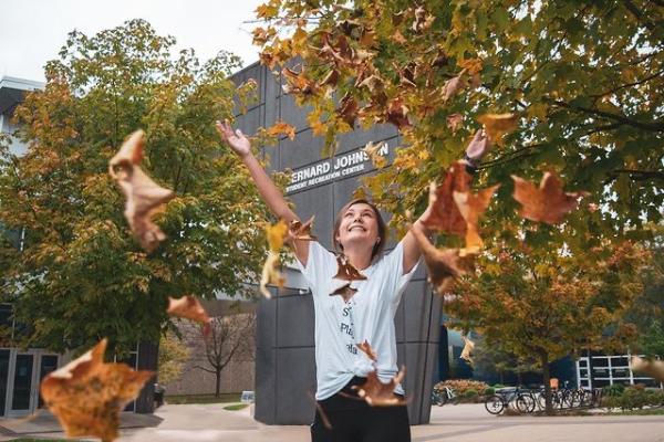A student stands in front of the two trees and UK Johnson Center and tosses fall-colored leaves into the air.