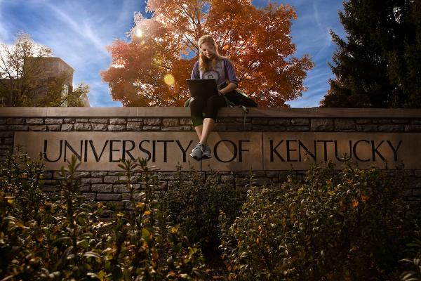 Stone "University of Kentucky" sign near the Administration Loop of Limestone. A student sits with their laptop, the Fall sunshine behind them.