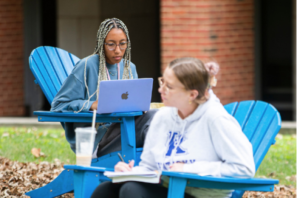 Two students sit outside sitting in blue Adirondack chairs. The student in the background is on their laptop. The student in the foreground is writing on their notepad.