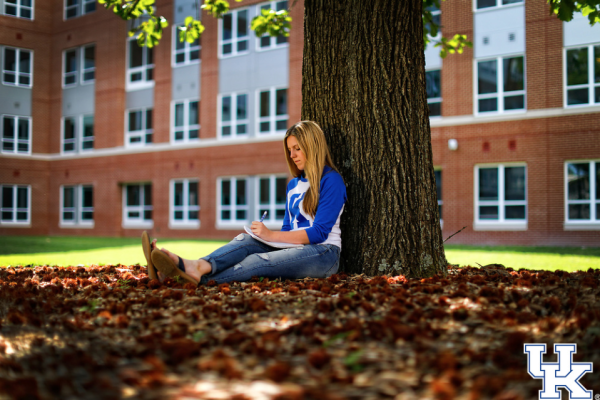 A student with long blonde hair sits under a tree in front of a red brick building and writes in a notebook.