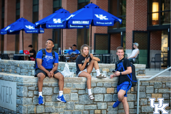 Three students sit on a brick wall outside of the UK student center. Other students sit on the patio at tables underneath UK-branded blue umbrellas.
