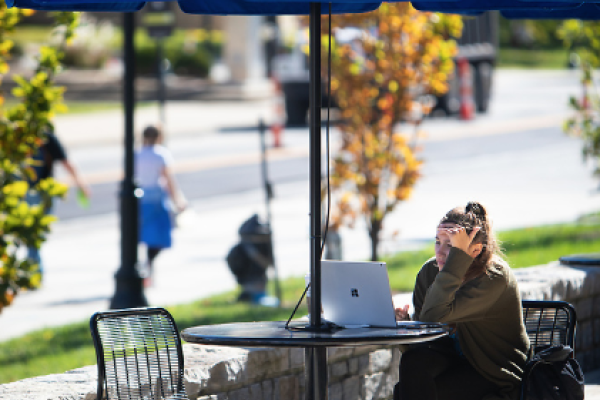 A student looks at their laptop as they sit underneath a UK-branded blue umbrella at an outdoor table. Their service dog looks at the camera.
