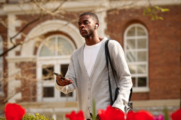 A student walking outside holds their phone out and glances at it. The foreground contains a flowerbed and in the background is a brick building.
