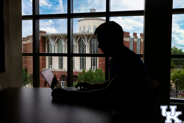 A student sits at a table cast in a shadow. The Willy T Young Library is in the background in the sun.