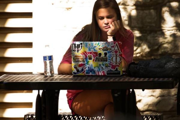 A student with long dark hair sits at a outdoor table and gazes at their heavily stickered laptop with an expression of deep concentration on their face.
