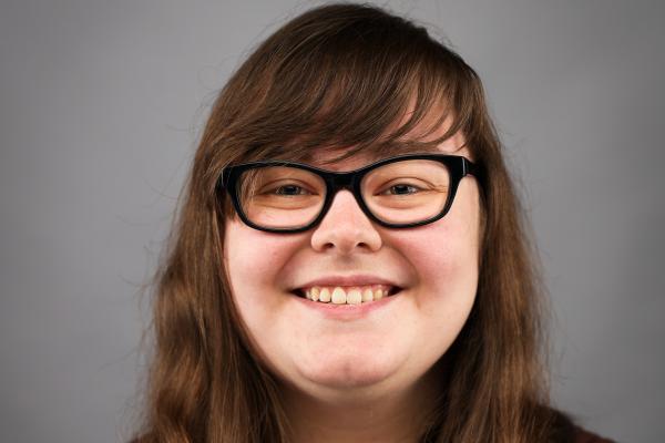 Headshot of a person with long brown hair, wearing black rimmed glasses.