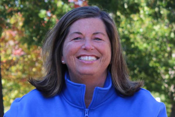 A woman with brown hair and a bright blue jacket sits smiling