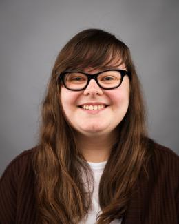 Headshot of a person with long brown hair with side swept bangs, wearing black rimmed glasses and a black cardigan with a white undershirt.