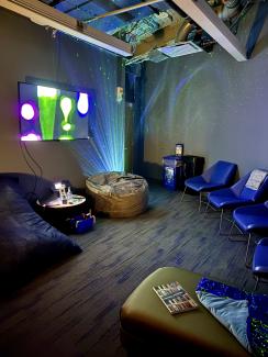 A dimly lit room with glowing vanity lights, a bean bag, neon projections, and a television displaying bright abstract shapes.