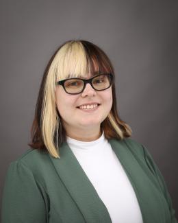 Professional headshot of a female with short calico style hair, wearing dark rimmed glasses and a green blazer with white shirt underneath.