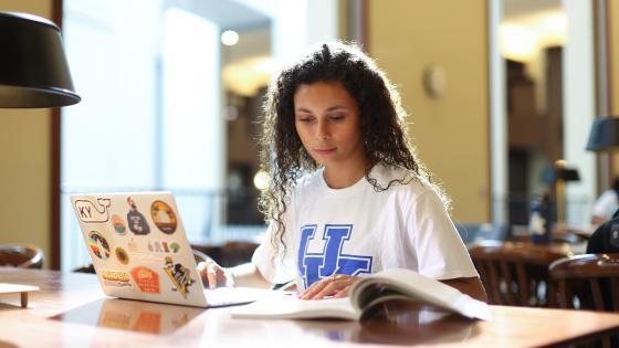 A student studies in the library common space with a book open on their right and laptop open on their left.