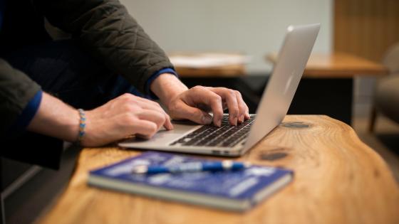 The torso of an individual is visible as they work on their laptop. A blue book and pen sit on the top of the wooden table next to them.