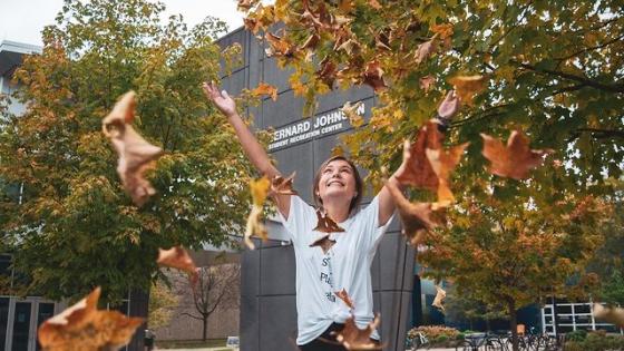 A student stands in front of the two trees and UK Johnson Center and tosses fall-colored leaves into the air.