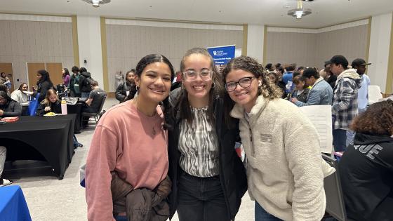 Three young women stand close together, smiling at the camera in a crowded indoor event space. They are holding papers and wearing casual clothing, including jackets and sweaters. The background features tables, booths, and other attendees engaged in conversations and activities.