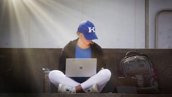 A person sitting criss crossed with a laptop wearing a blue UK baseball cap.