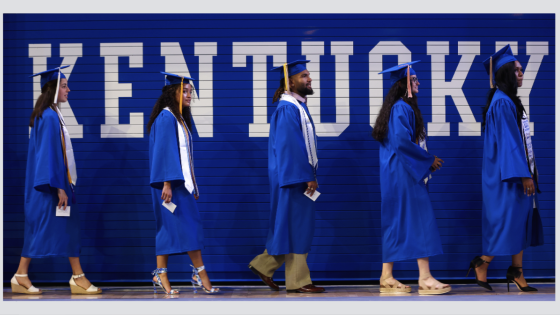 Students walking in a line wearing graduation attire with the word Kentucky on the wall behind them.