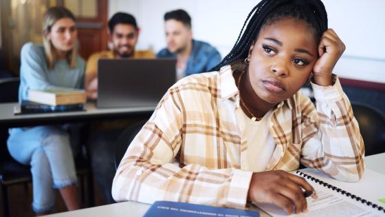 A person in a plaid shirt appears thoughtful at a desk in a classroom, with three other students and a laptop in the background.