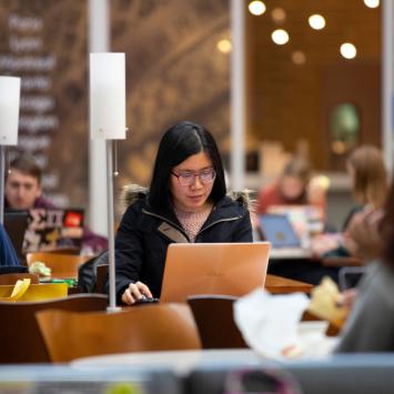 A student glances at their laptop with a look of concentration as they sit at a table underneath a white lamp. Students surround them at other tables in various stages of lounging and concentration.