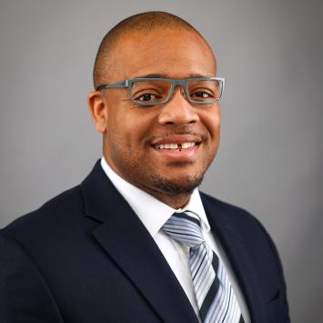 A professional headshot of a smiling male wearing a navy blue suit, white dress shirt, and a striped tie. He has short hair, a well-groomed beard, and wears gray-framed glasses. The background is a neutral gray color.