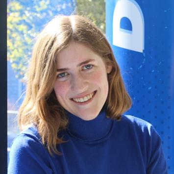 A young woman with red hair and a blue sweater sits and smiles