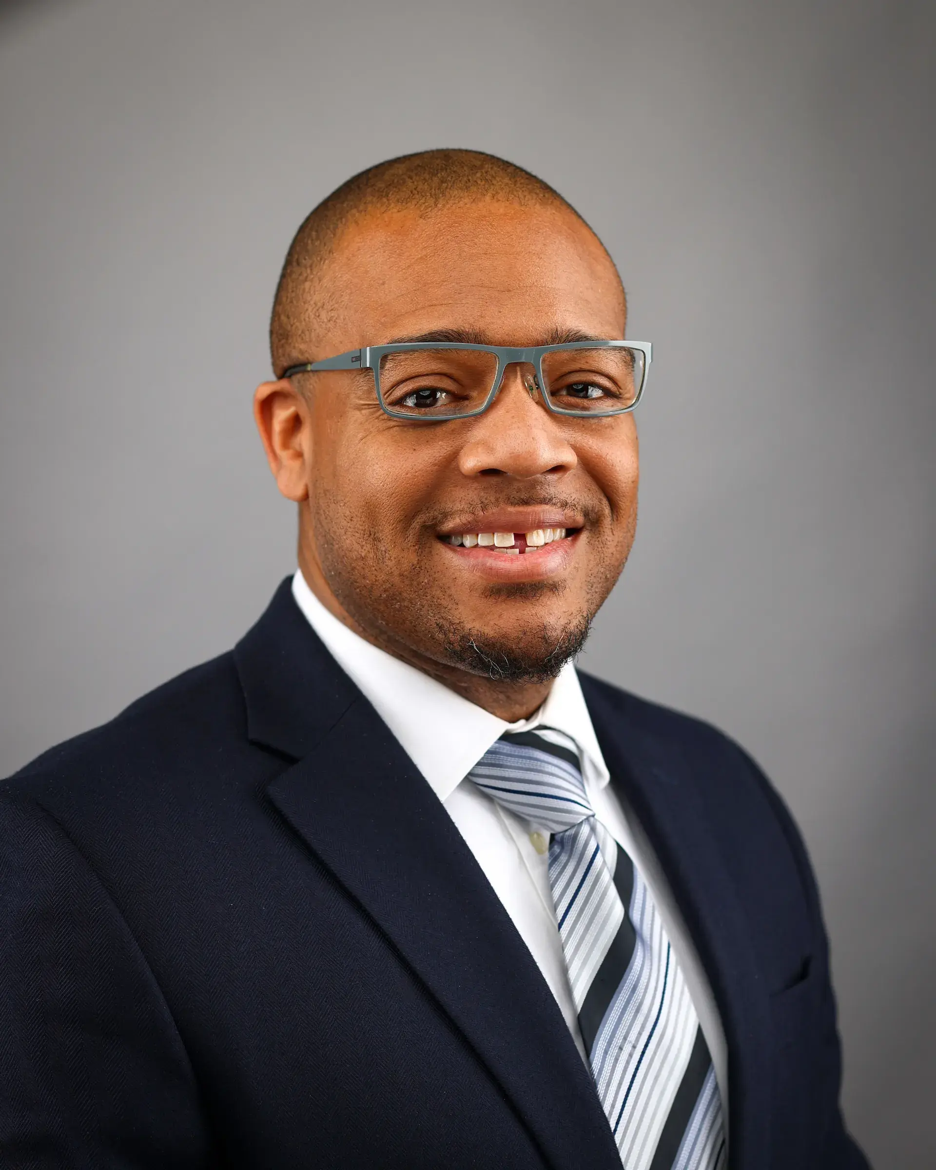 A professional headshot of a smiling male wearing a navy blue suit, white dress shirt, and a striped tie. He has short hair, a well-groomed beard, and wears gray-framed glasses. The background is a neutral gray color.