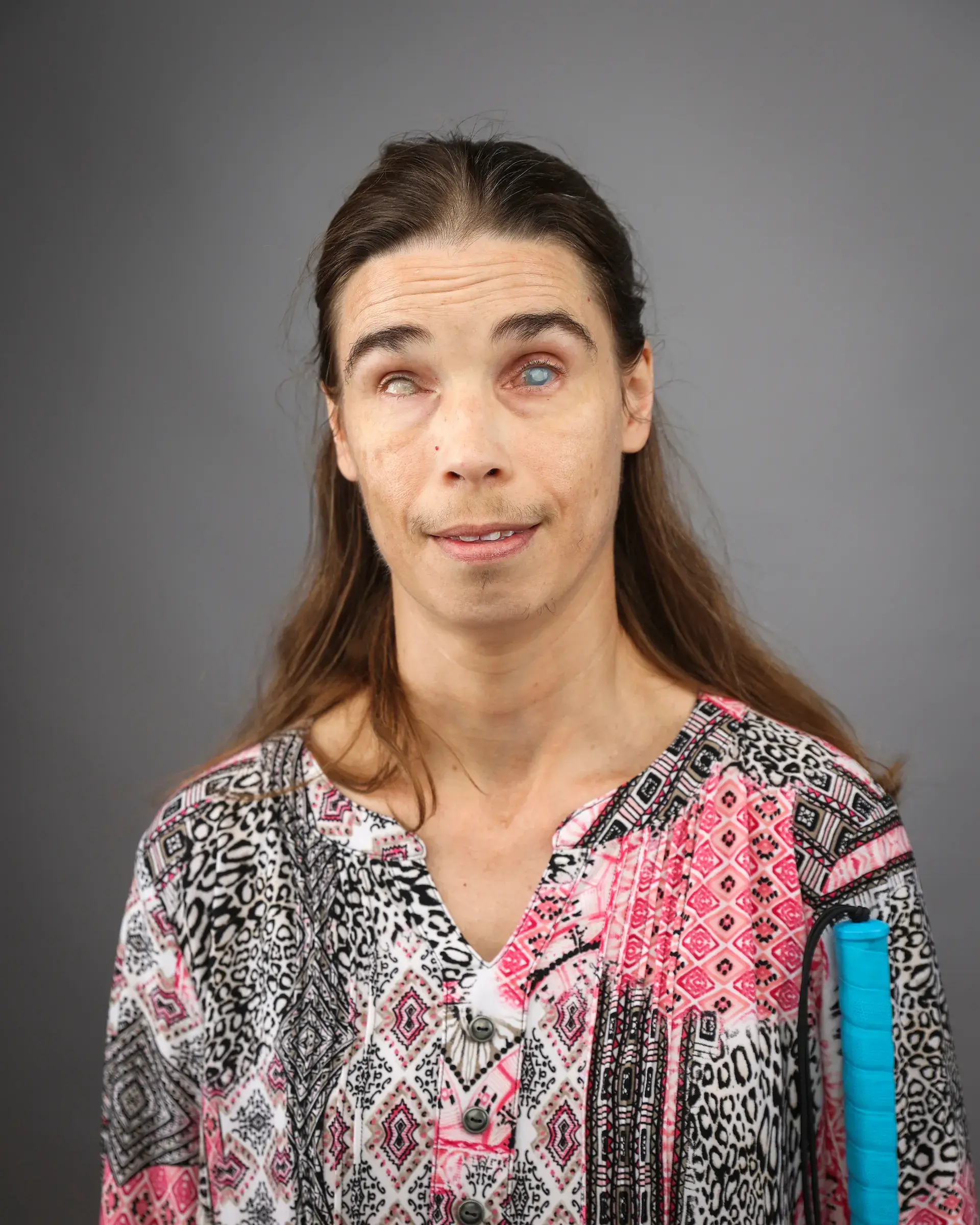 A woman with medium length brown hair wearing a colorful pink and black v-neck top. A white cain is visible.