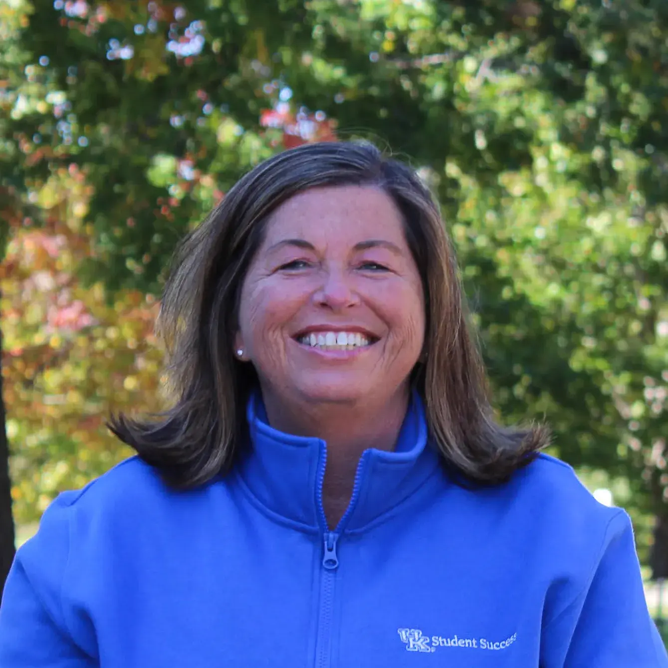 A woman with brown hair and a bright blue jacket sits smiling