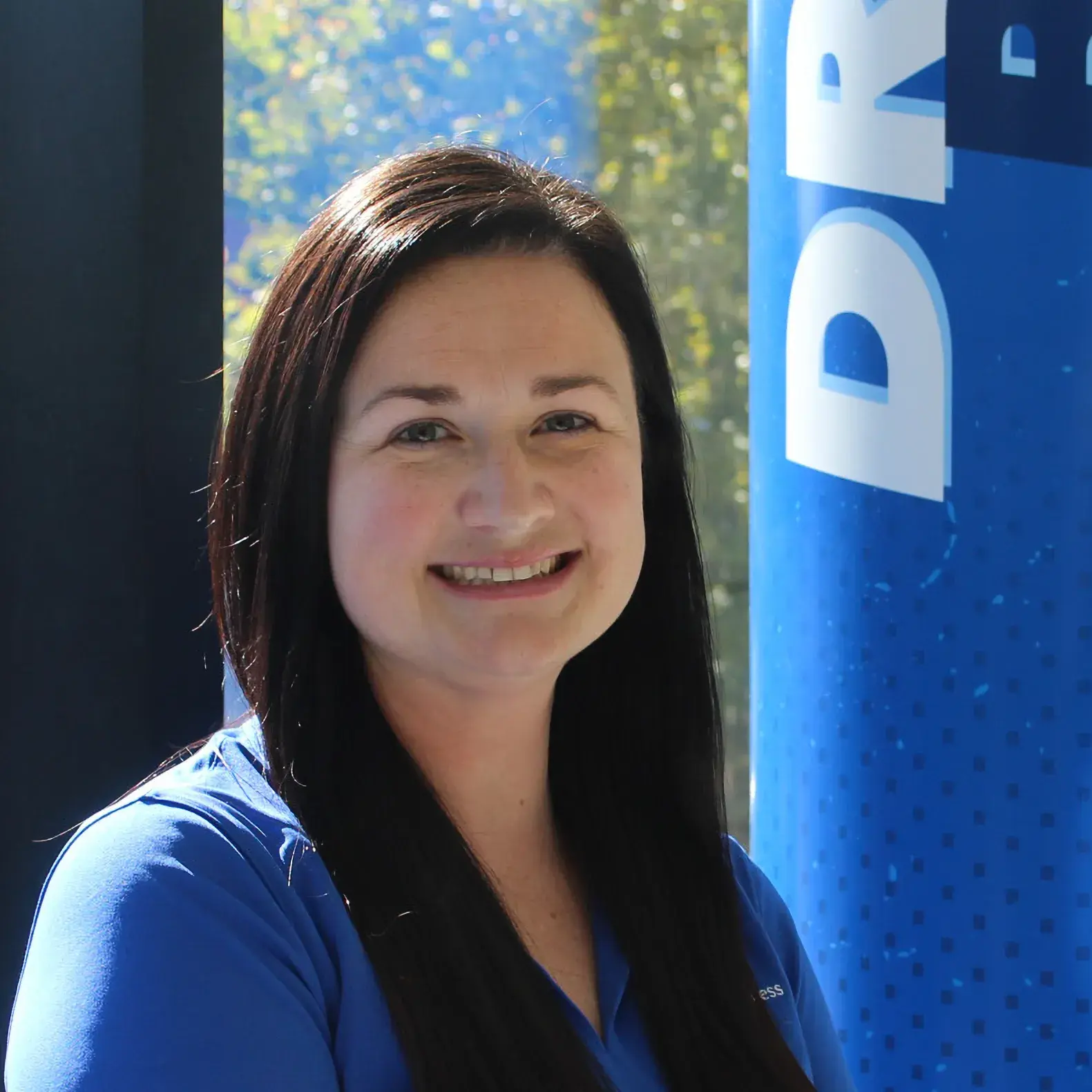 A woman with dark brown hair and a blue shirt sits smiling